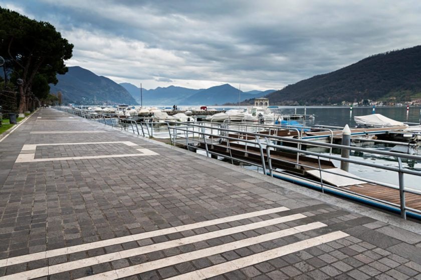 a walkway by a lake with yachts and mountains in the background