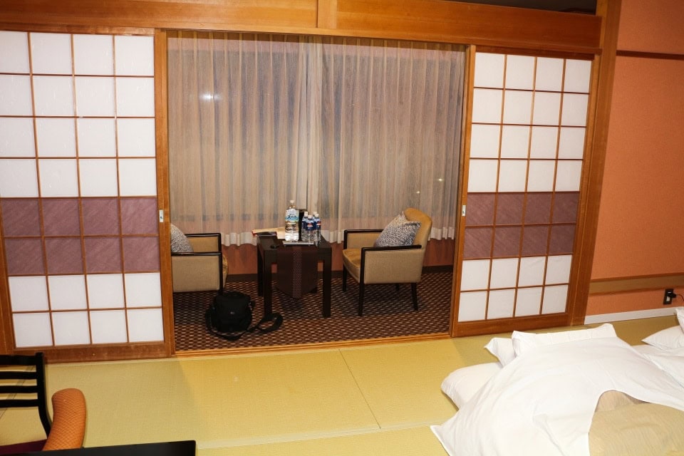 relaxation area in a ryokan in japan with chairs, table, and waters, tatami mat floor and futon beds visible as well