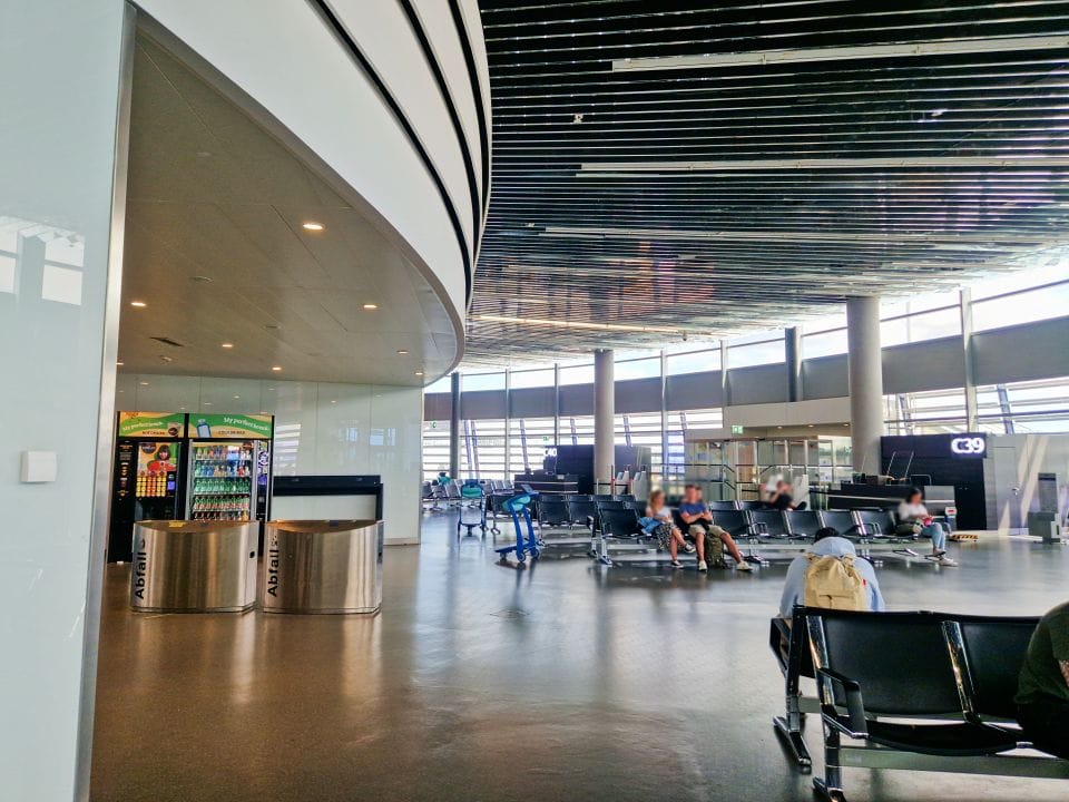 waiting for a flight in an airport lobby - there are people sitting on chairs, vending machines, and trash cans visible